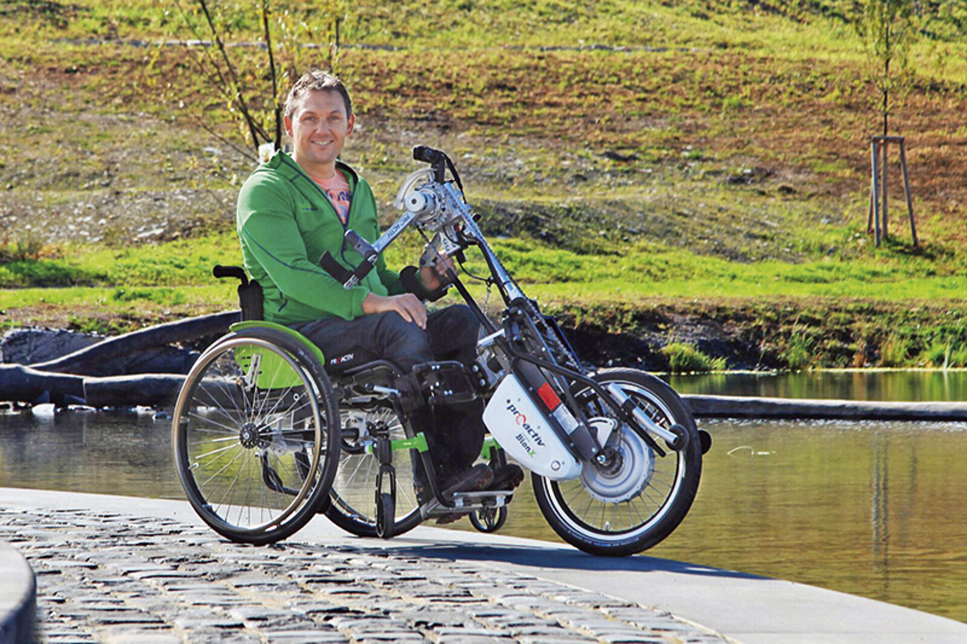 Ein Mann sitzt in einem Rollstuhl mit elektrischem Zuggerät am Rand eines Gewässers und lacht in die Kamera.