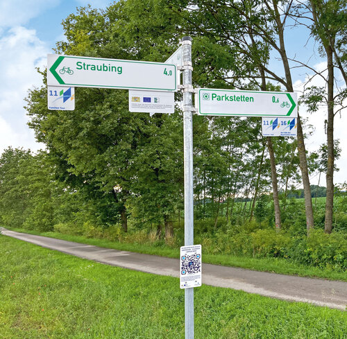 Ein Radweg wird begrenzt einerseits durch eine Wiese, auf der anderen Seite durch Bäume und Sträucher. Die Radwegschilder weisen gegenüberliegend die Richtungen nach Straubing und Parkstetten.